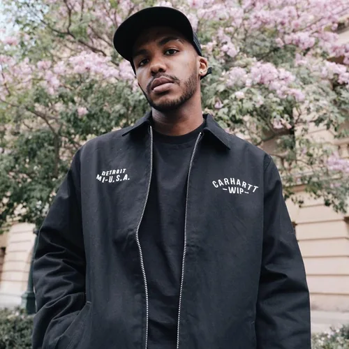 Danyon in a black cap and dark work jacket, standing in front of a blossoming tree